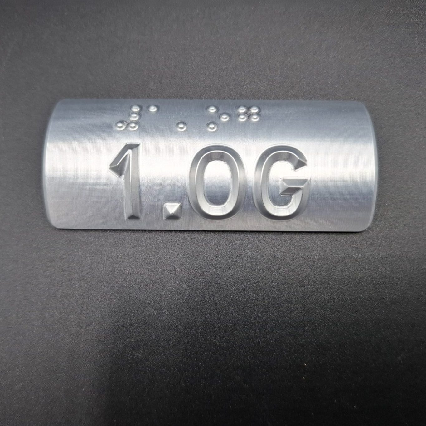 Silberne Plakette mit der Zahl "1.0G" und Brailleschrift darunter.