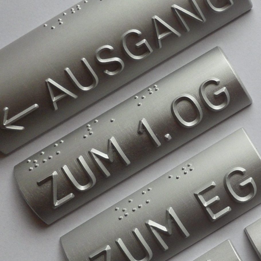Metallschilder mit Braille und erhabenen Buchstaben als Wegweiser.