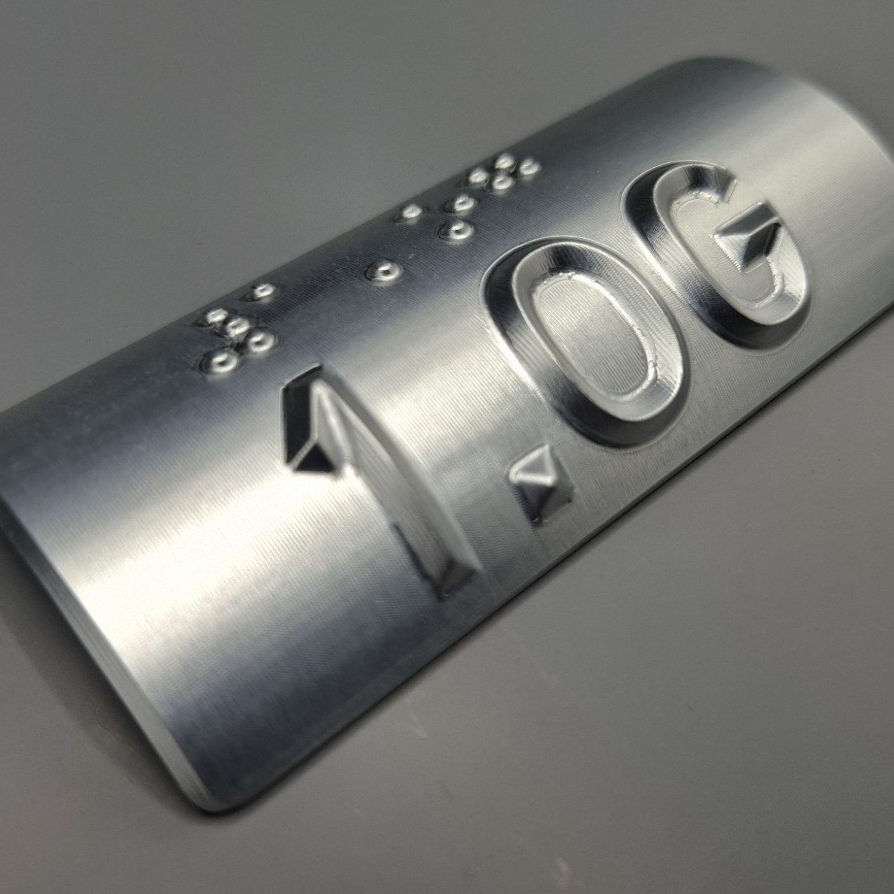 Metallplatte mit erhabener Zahl "1.0G" und Braille-Schrift.